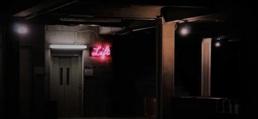 Entrada oscura con un cartel de neón rosa que indica “Lift”, en un ambiente industrial y alternativo