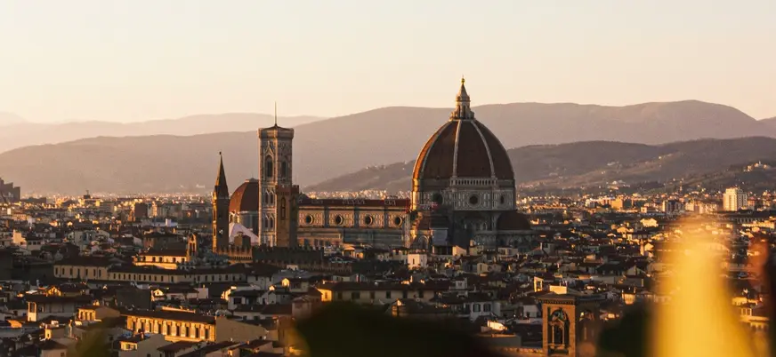 Vista panorámica de Florencia, Italia, al atardecer, destacando la Catedral de Santa María del Fiore con su cúpula de Brunelleschi y la torre del campanario, rodeadas de edificios históricos y montañas al fondo.