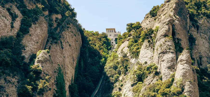 Funicular de Sant Joan subiendo por la grieta de roca de Montserrat, rodeado de vegetación y edificio en la cima.