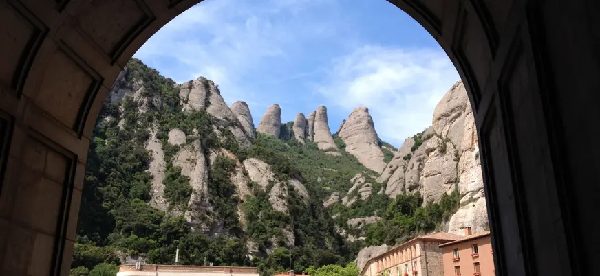 Vista enmarcada por un arco desde el monasterio de Montserrat hacia los picos rocosos, cubiertos de vegetación.