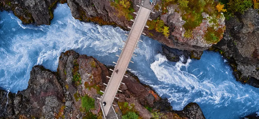 Puente sobre las aguas turquesas del río en Hraunfossar, Islandia, rodeado de rocas volcánicas y vegetación.
