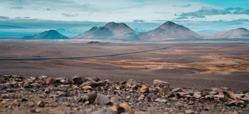 Paisaje volcánico cerca de Egilsstaðir, Islandia, con montañas y carretera entre terrenos áridos bajo cielo azul.
