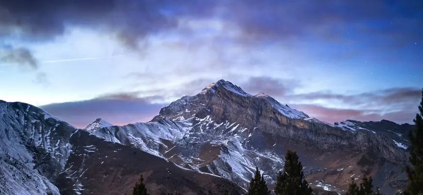Paisaje nocturno de montaña nevada bajo un cielo estrellado y nublado en Aínsa.