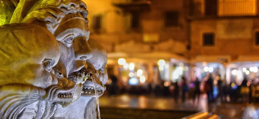 Primer plano de escultura en fuente romana con plaza iluminada al fondo de noche.