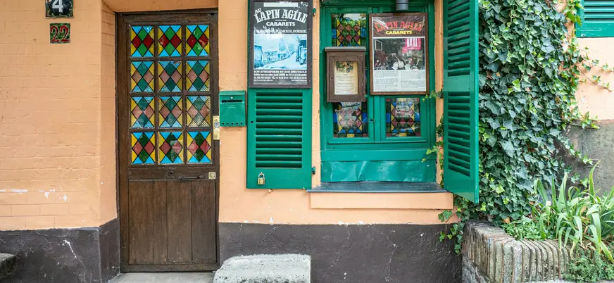 Entrada decorada de un bar en Montmartre, París, con objetos artísticos colgados en la pared, puerta ornamentada y macetas en una calle empedrada bajo luz natural.