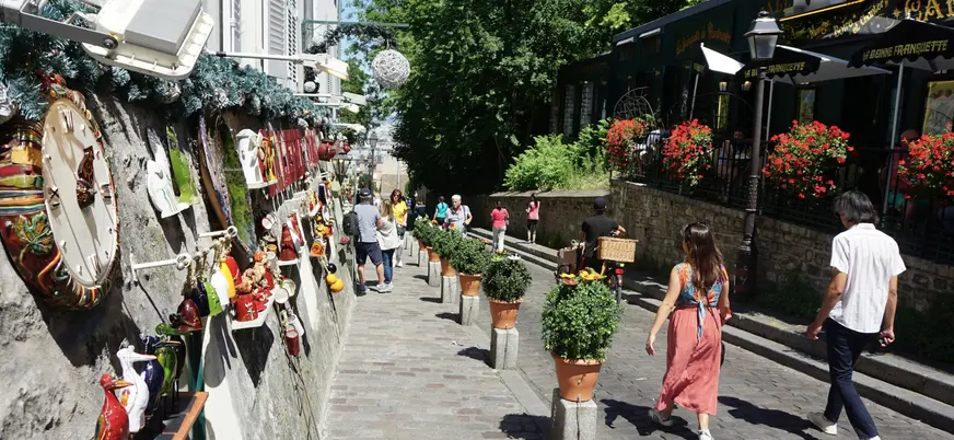 Calle empedrada en Montmartre, París, decorada con macetas y objetos artísticos en las paredes, con personas paseando bajo luz natural y vegetación al fondo.