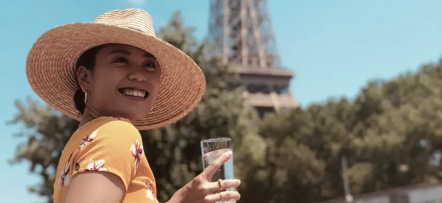 Mujer sonriente con sombrero y bebida frente a la Torre Eiffel en un día soleado.