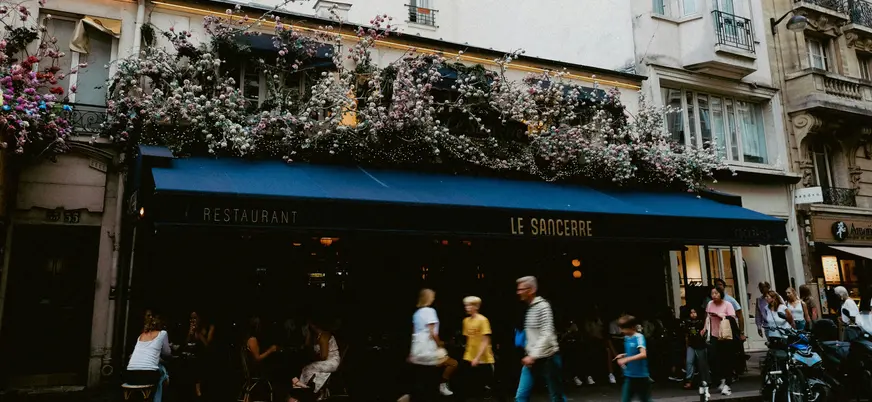 Fachada del restaurante Le Sancerre en París, con toldo azul oscuro y decoración floral abundante en el balcón superior, mientras varias personas caminan por la acera.