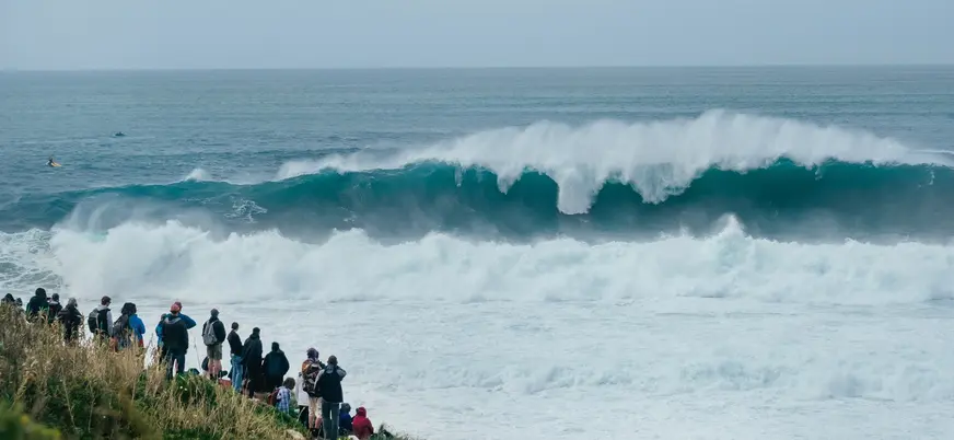 Turistas observando una ola gigante rompiendo en la costa de Nazaré, Portugal.