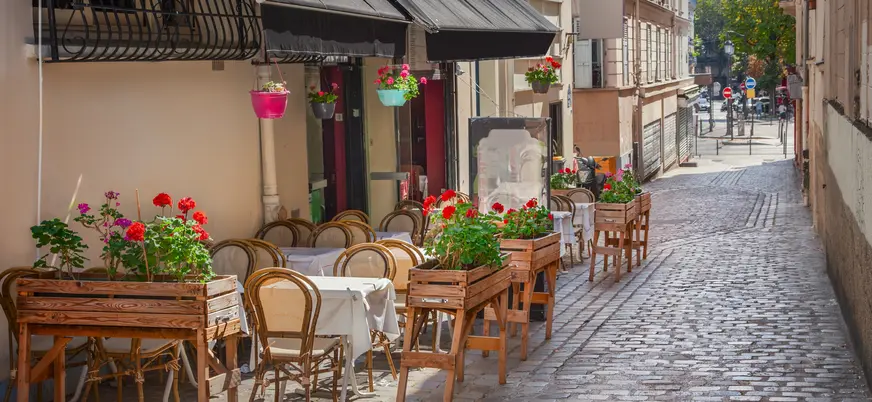Calle empedrada en Montmartre, París, con mesas y sillas de madera decoradas con flores frente a un café, bajo luz natural en un entorno bohemio.