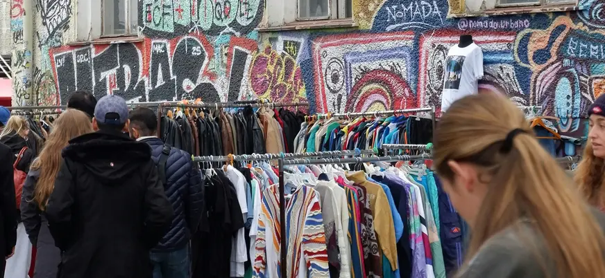 Gente revisando percheros de ropa con grafiti al fondo en Berlín.