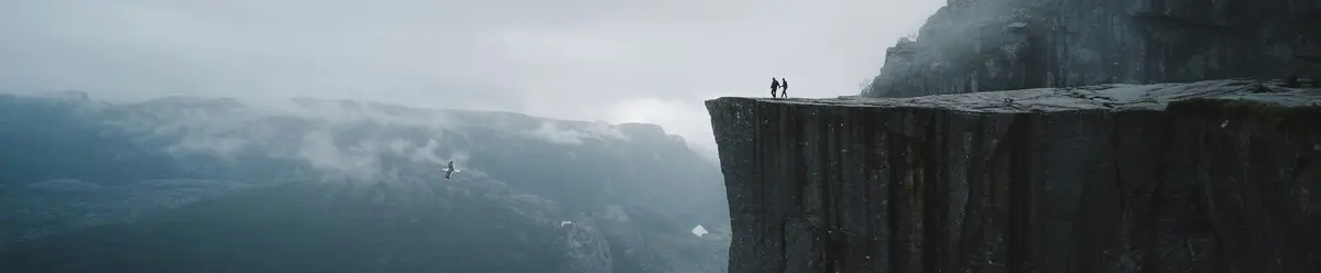 Dos personas en la cima de Preikestolen (Pulpit Rock) sobre el fiordo Lysefjord, Noruega, con niebla y nubes.