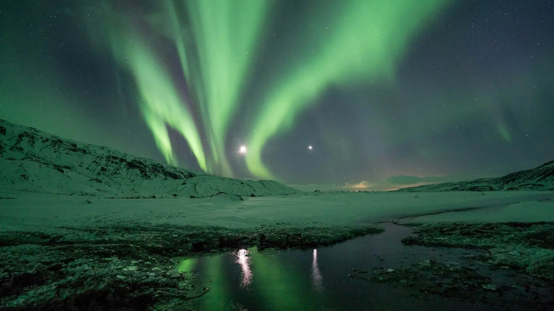 Aurora boreal sobre un paisaje nevado en Islandia, con reflejos verdes iluminando el cielo y las montañas.