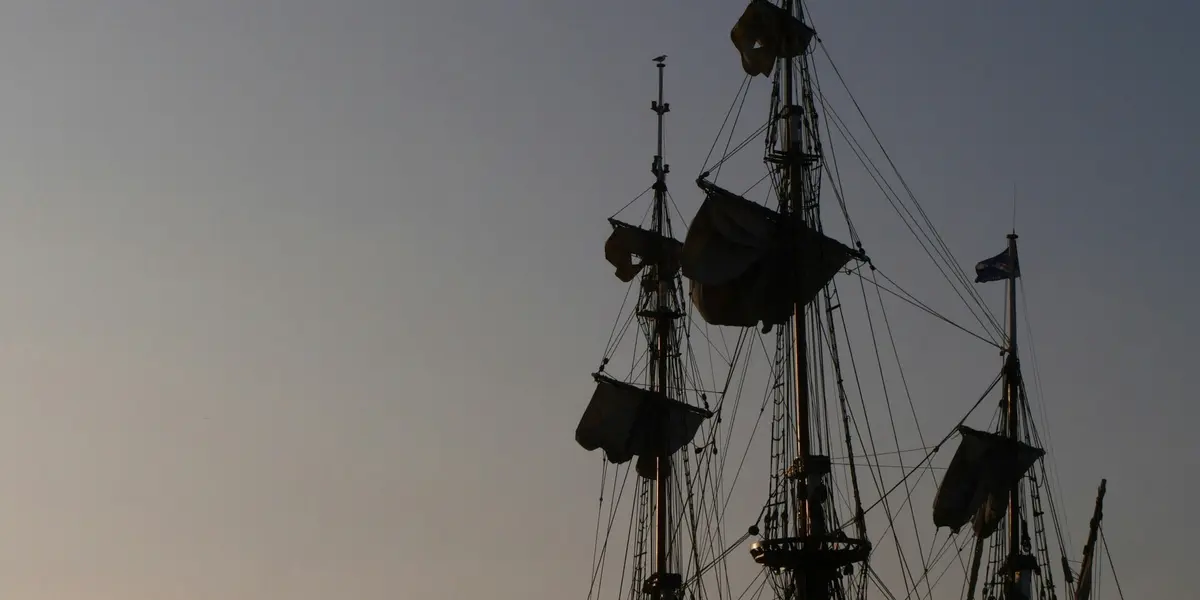 Mástiles y velas de un barco antiguo o réplica al atardecer, siluetas oscuras contra un cielo despejado