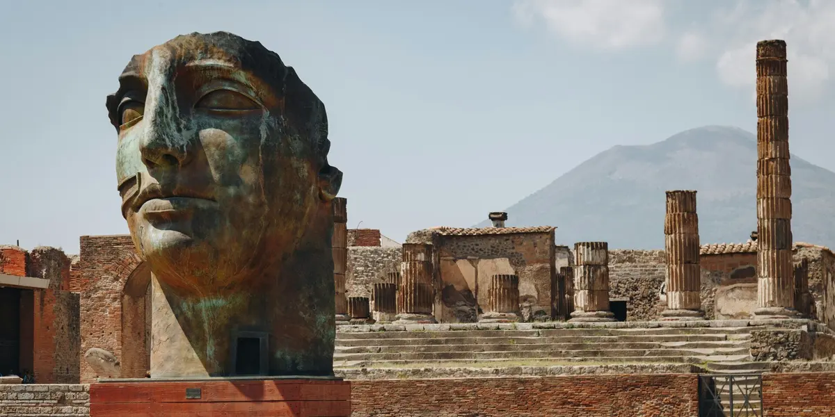 Cabeza de bronce en Pompeya con el Monte Vesubio y las ruinas de columnas romanas de fondo.