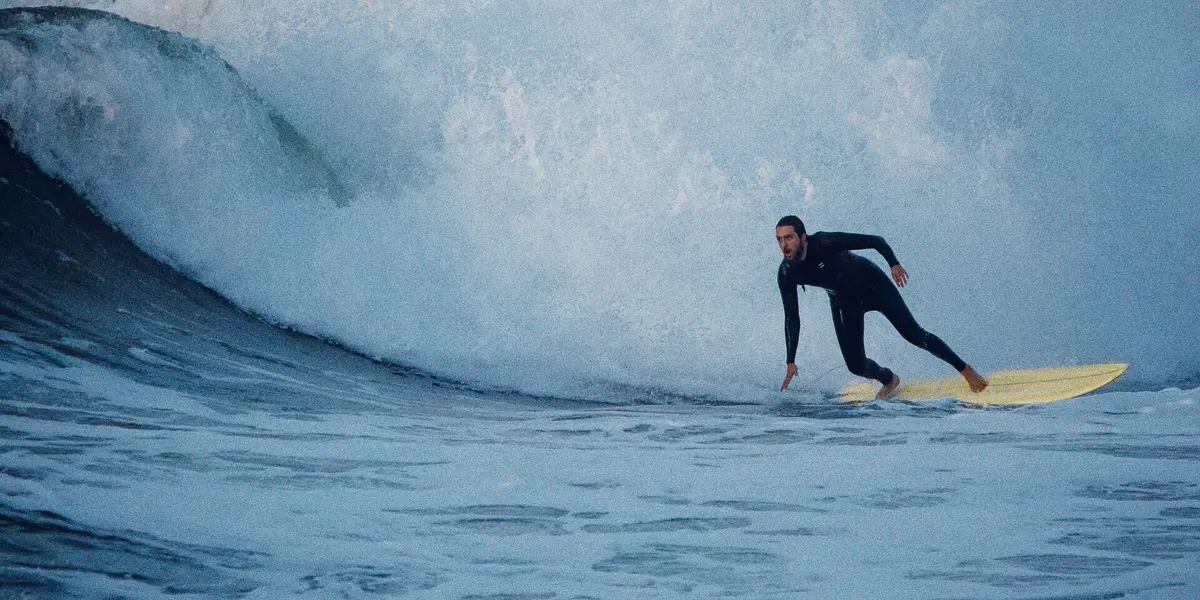 Surfista en traje de neopreno montando una tabla amarilla sobre una gran ola blanca y azul.