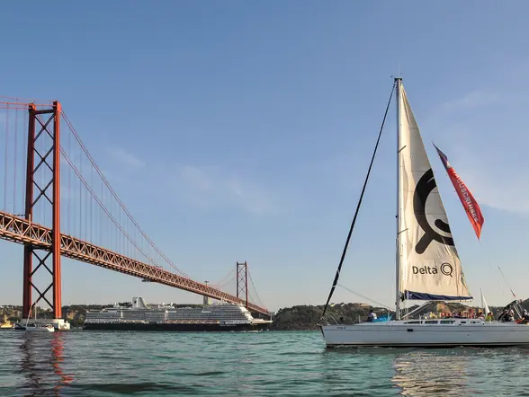 Velero navegando por el río Tajo con el puente 25 de Abril al fondo y un crucero cerca de la orilla, bajo un cielo despejado.