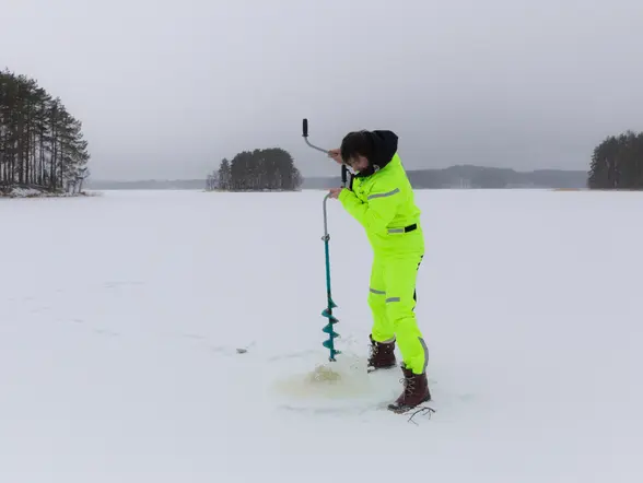 Persona perforando el hielo con un taladro manual en un lago congelado, vestida con traje térmico color neón, en un paisaje invernal rodeado de árboles.