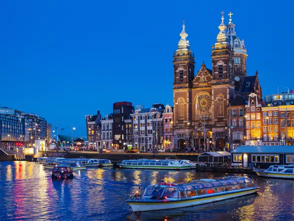 Vista nocturna de los canales de Ámsterdam con barcos turísticos navegando, edificios históricos iluminados y la iglesia de San Nicolás destacando en el fondo.