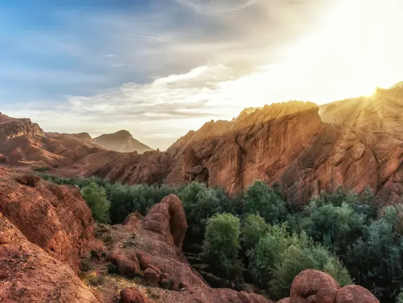 Paisaje del Alto Atlas al atardecer con formaciones rocosas rojizas y un oasis de vegetación frondosa entre las montañas.