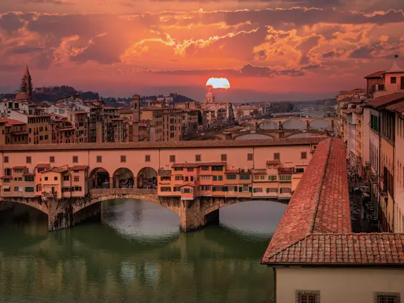 Vista del Ponte Vecchio en Florencia al atardecer, con el sol poniéndose sobre el río Arno y edificios históricos iluminados por tonos cálidos.