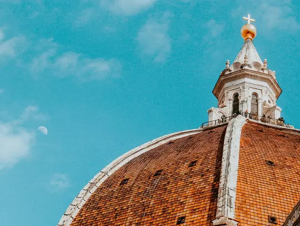 Detalle de la cúpula de la Catedral de Santa María del Fiore en Florencia, conocida como el Duomo, con tejas rojizas y linterna coronada por una cruz dorada sobre un cielo azul.