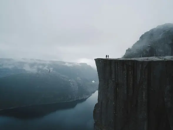 Dos personas en la cima de Preikestolen (Pulpit Rock) sobre el fiordo Lysefjord, Noruega, con niebla y nubes.