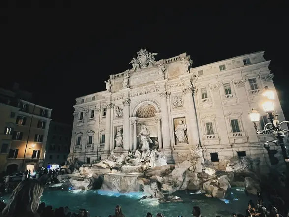 La Fontana di Trevi iluminada de noche con visitantes en la plaza de Roma.