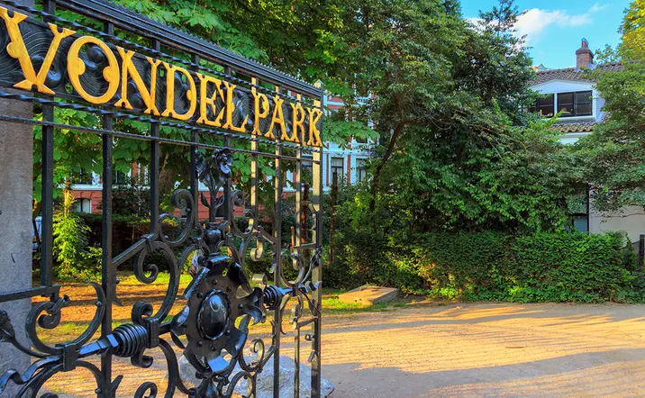 Puerta de hierro forjado con el letrero "Vondelpark" en amarillo, entrada al famoso parque rodeada de árboles y casas residenciales en Ámsterdam.