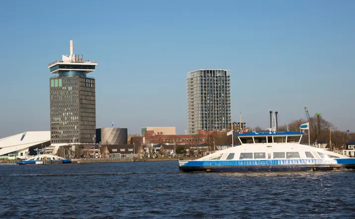 Ferry blanco y azul navegando por el río IJ en Ámsterdam, con el edificio A'DAM Tower, el museo Eye y otros edificios modernos.