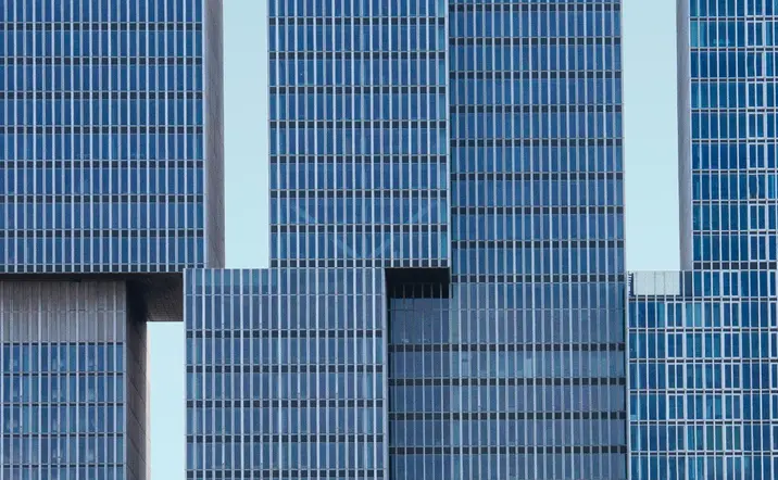 Fachada de un moderno edificio de oficinas compuesto por varios bloques rectangulares de vidrio azul, alineados de forma asimétrica, sobre un cielo despejado de color celeste.