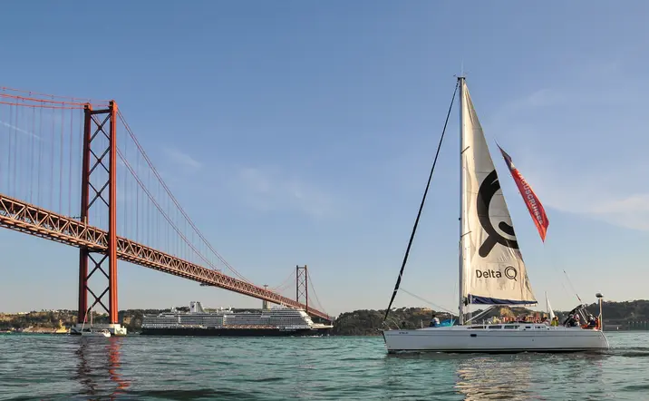 Velero navegando por el río Tajo con el puente 25 de Abril al fondo y un crucero cerca de la orilla, bajo un cielo despejado.