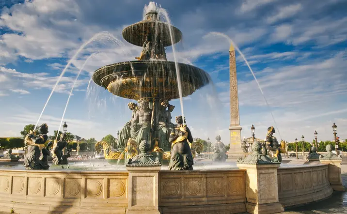 Fuente histórica en París con estructura de piedra y detalles arquitectónicos rodeada de vegetación.