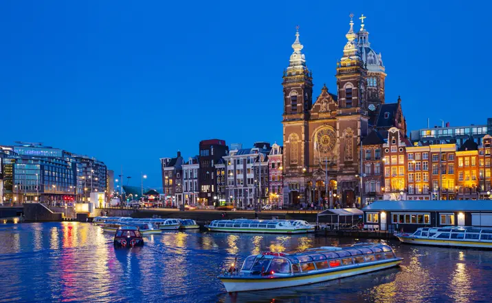 Vista nocturna de los canales de Ámsterdam con barcos turísticos navegando, edificios históricos iluminados y la iglesia de San Nicolás destacando en el fondo.