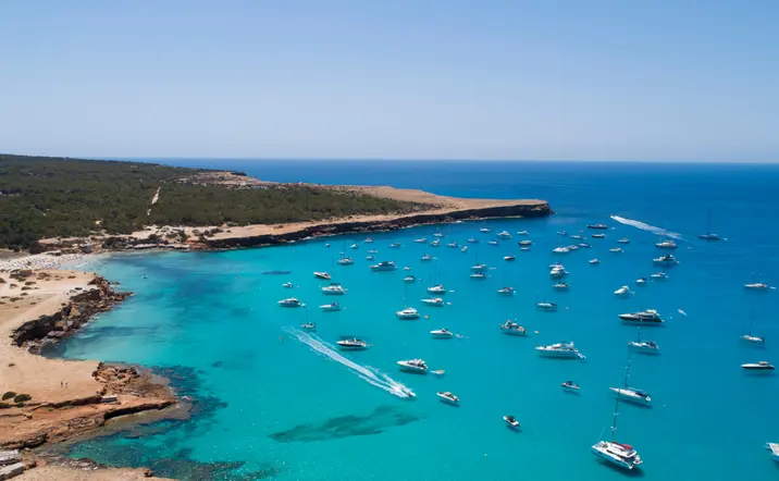 Cala Saona vista desde lo alto, con barcos, vegetación y aguas de un azul intenso.