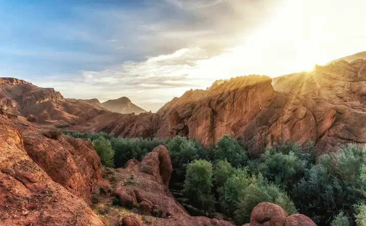 Paisaje del Alto Atlas al atardecer con formaciones rocosas rojizas y un oasis de vegetación frondosa entre las montañas.