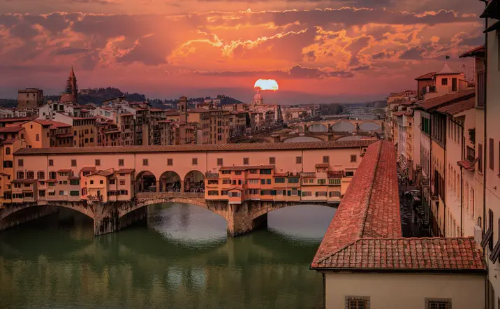 Vista del Ponte Vecchio en Florencia al atardecer, con el sol poniéndose sobre el río Arno y edificios históricos iluminados por tonos cálidos.