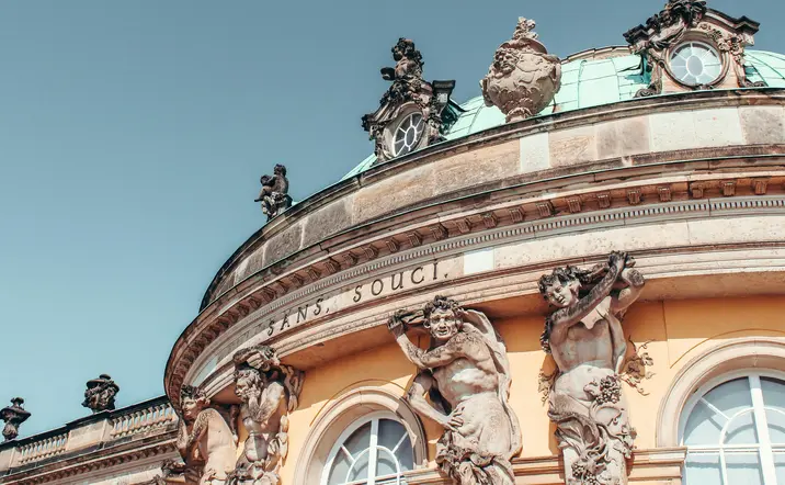 Detalle de la fachada del Palacio de Sanssouci en Potsdam, con estatuas de atlantes, inscripciones y la cúpula verde.