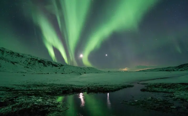 Aurora boreal sobre un paisaje nevado en Islandia, con reflejos verdes iluminando el cielo y las montañas.