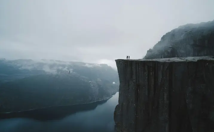 Dos personas en la cima de Preikestolen (Pulpit Rock) sobre el fiordo Lysefjord, Noruega, con niebla y nubes.