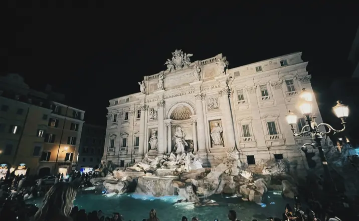 La Fontana di Trevi iluminada de noche con visitantes en la plaza de Roma.